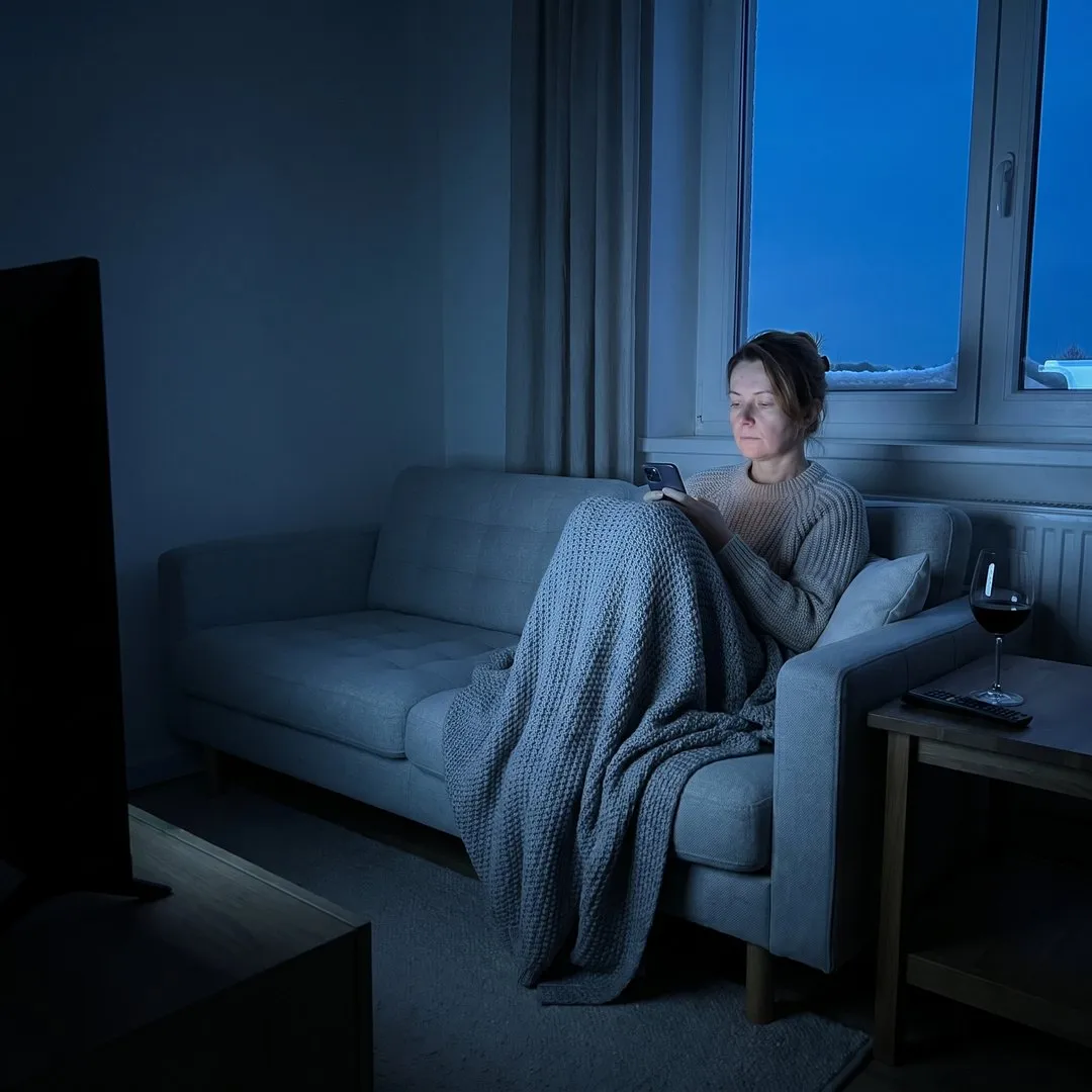 Frau sitzt im Dezember abends allein unter einer Decke auf dem Sofa und schaut aufs Handy, daneben ein Glas Rotwein, draußen Schnee auf dem Fensterbrett