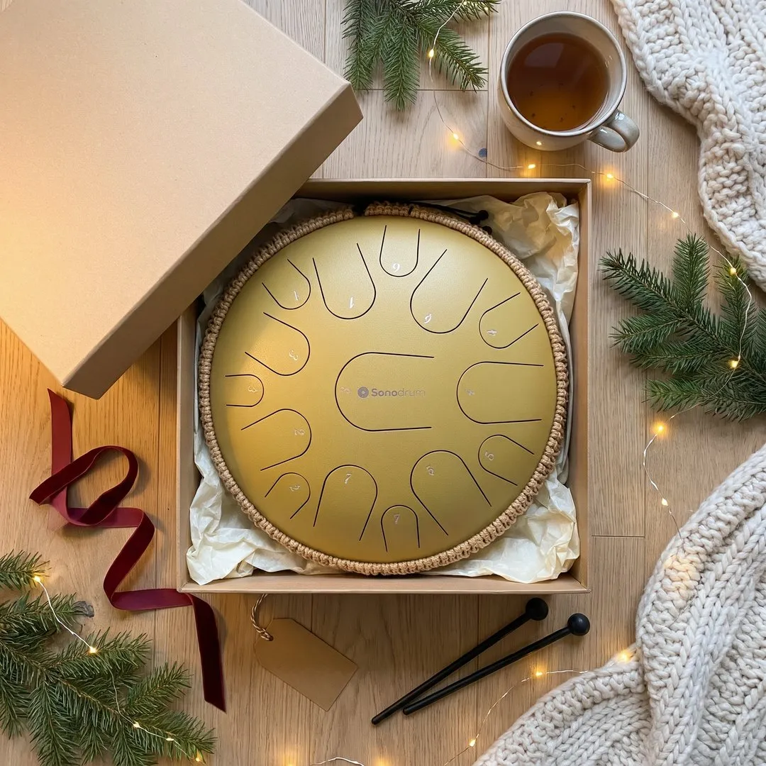 Goldene Sonodrum Zungentrommel in einer offenen Geschenkbox mit Seidenpapier, daneben Schlägel, Tannenzweige und eine Lichterkette